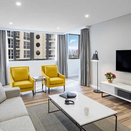 Meriton Suites Bondi Junction Sydney Bagian luar foto The photo shows a modern, well-lit living room. It features a light gray sofa with a textured fabric. There are two bright yellow armchairs positioned near a window, which allows natural light to enter the space. The room has light gray curtains and 