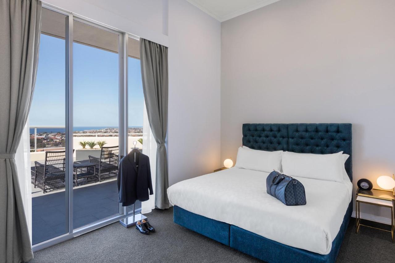 Meriton Suites Bondi Junction Sydney Bagian luar foto The photo shows a stylish bedroom featuring a large bed with a tufted blue headboard. There's a neatly organized outfit consisting of a suit hanging on a stand, along with shoes placed on the floor. The room has large windows that provide a view of t