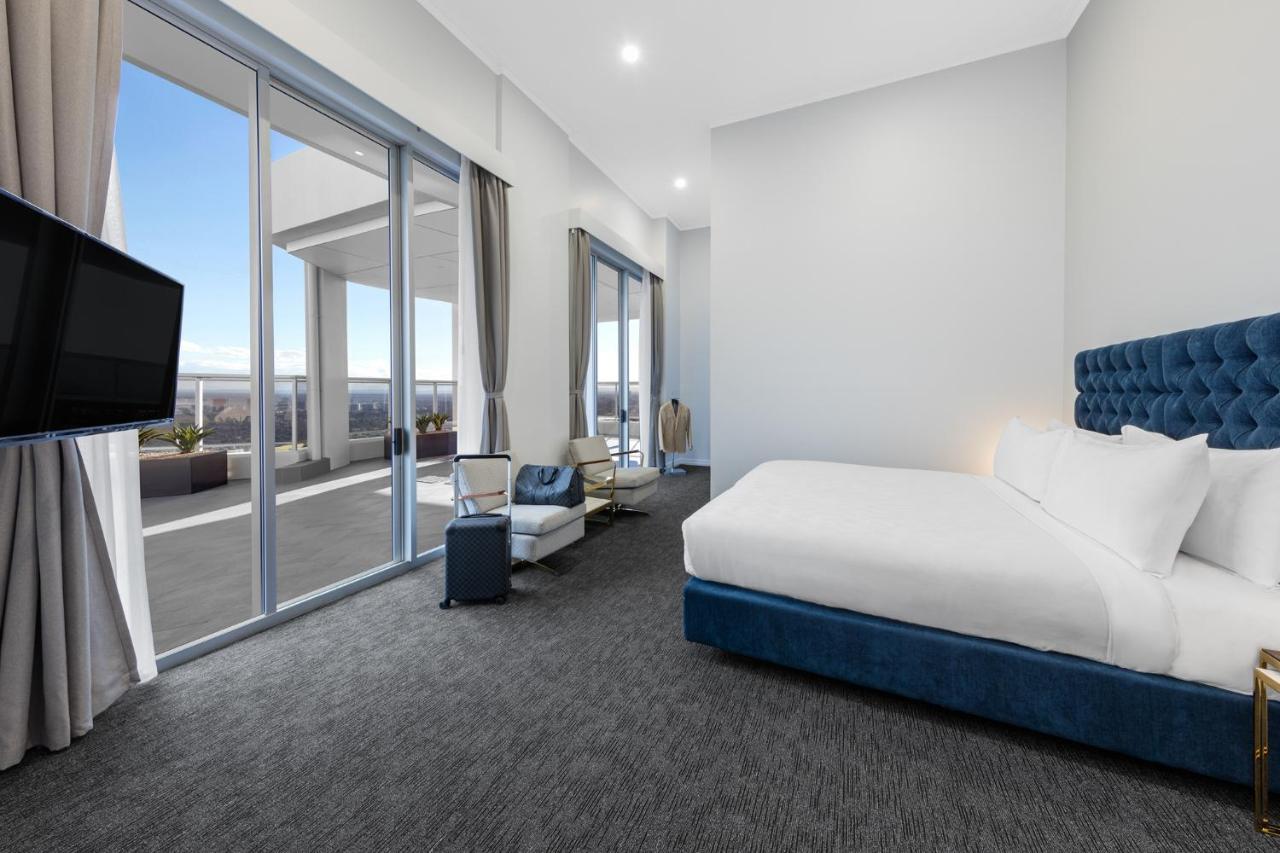 Meriton Suites Bondi Junction Sydney Bagian luar foto The photo shows a modern and spacious hotel room. It features a large bed with a blue headboard, a flat-screen TV mounted on the wall, and a cozy seating area with a chair and small table. The room has large windows that offer natural light and a vie