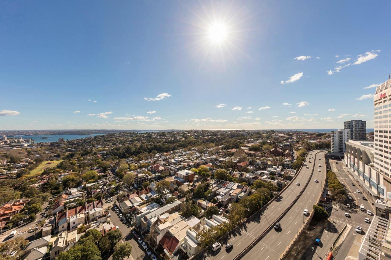 Meriton Suites Bondi Junction Sydney Bagian luar foto The photo shows a panoramic view of a city landscape under a clear blue sky. In the foreground, there is a road with vehicles traveling along it, bordered by trees and buildings. In the background, you can see a residential area with various houses, 