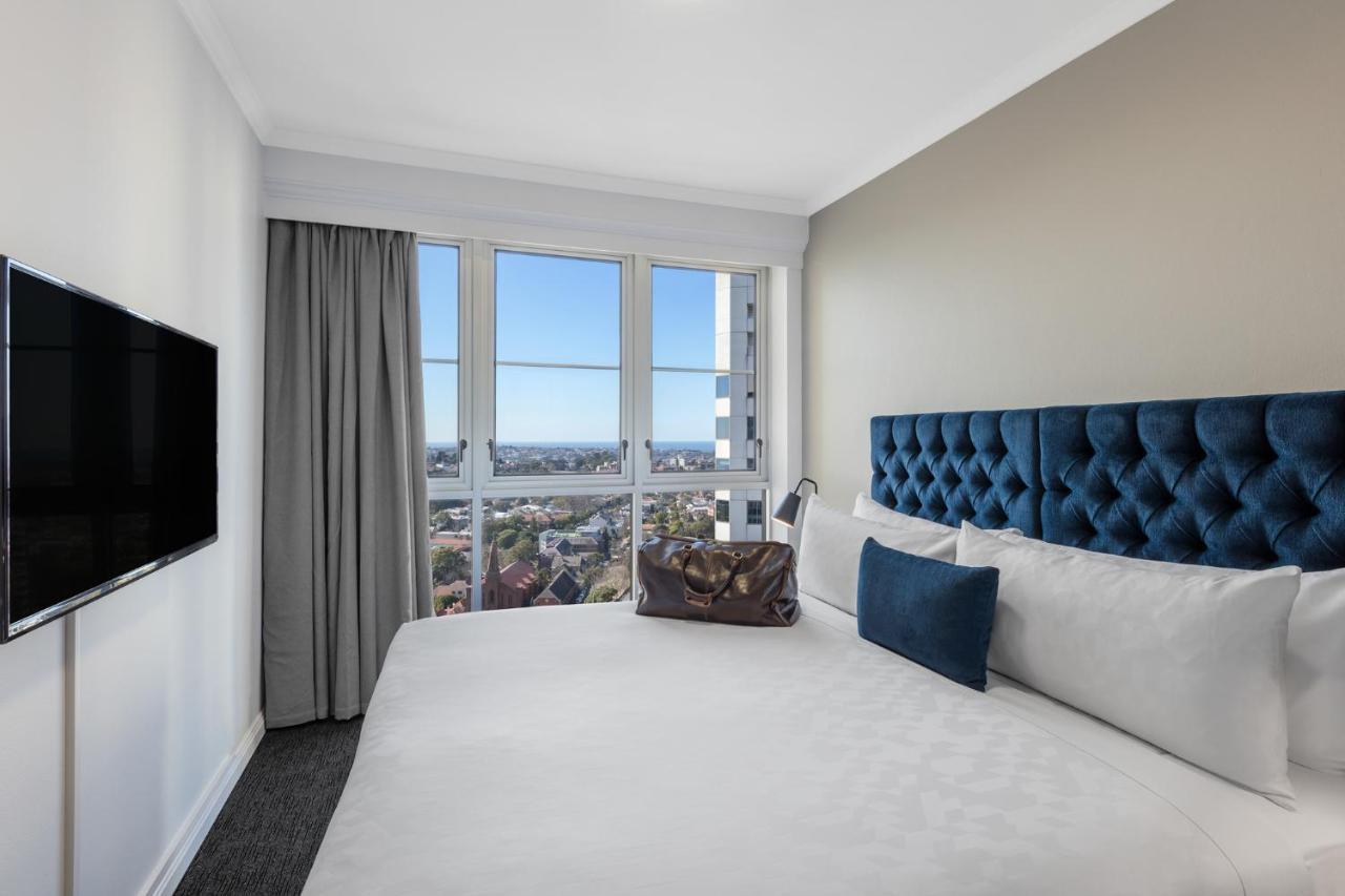 Meriton Suites Bondi Junction Sydney Bagian luar foto The photo shows a modern hotel room featuring a large bed with a tufted blue headboard. Beside the bed, there is a stylish bag placed on the bed. The room has large windows that provide a view of the outside, allowing natural light to fill the space.
