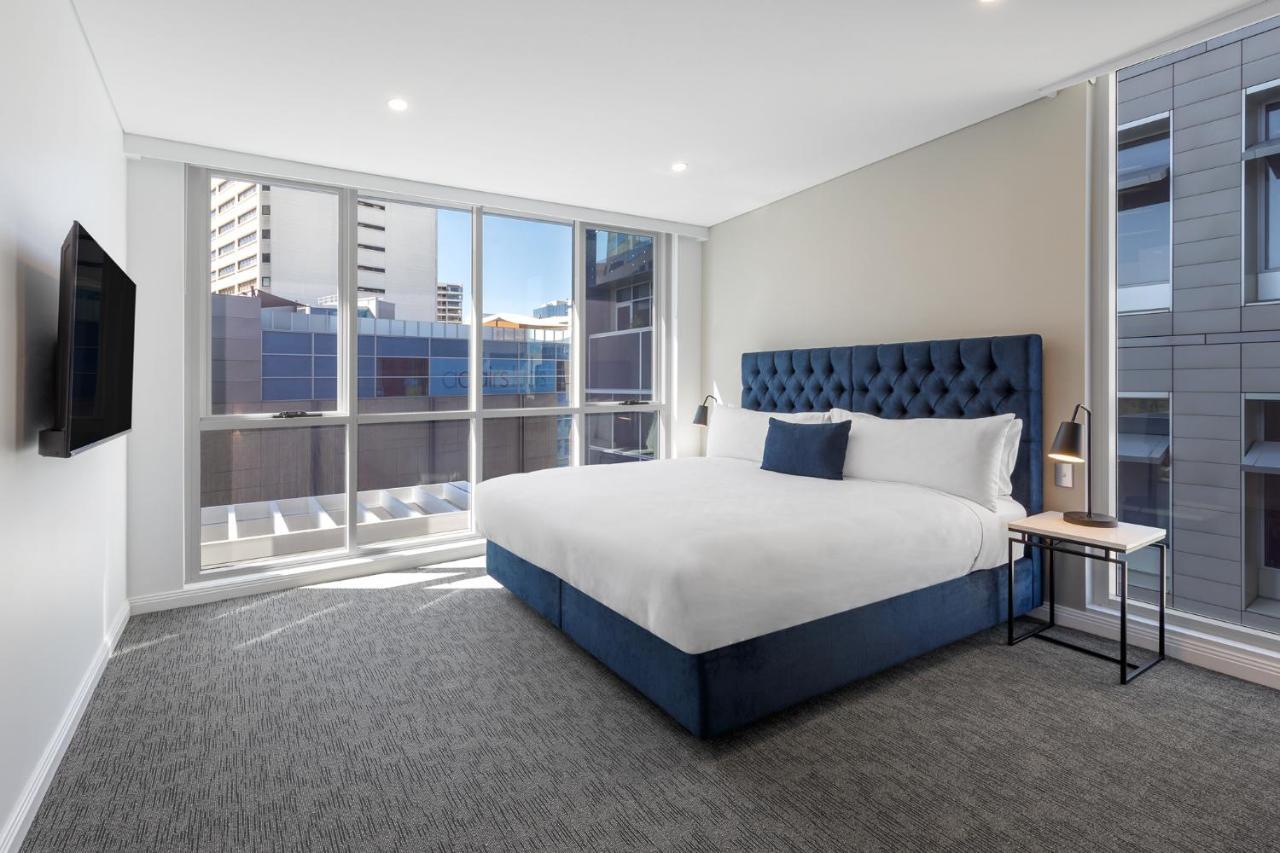 Meriton Suites Bondi Junction Sydney Bagian luar foto The image shows a modern hotel room with a large, comfortable bed featuring a blue upholstered headboard. The room has large windows that allow natural light to enter, providing a good view of the surrounding buildings. There is a small bedside table