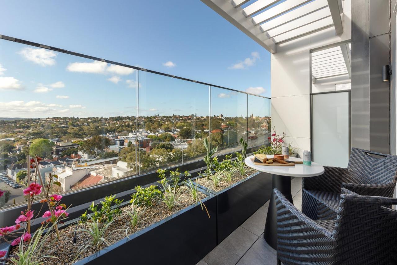 Meriton Suites Bondi Junction Sydney Bagian luar foto The photo shows a modern balcony with a glass railing, enhancing the view of the surrounding area, which includes various rooftops and greenery in the distance. There is a small round table set with a decorative floral arrangement and some items, lik