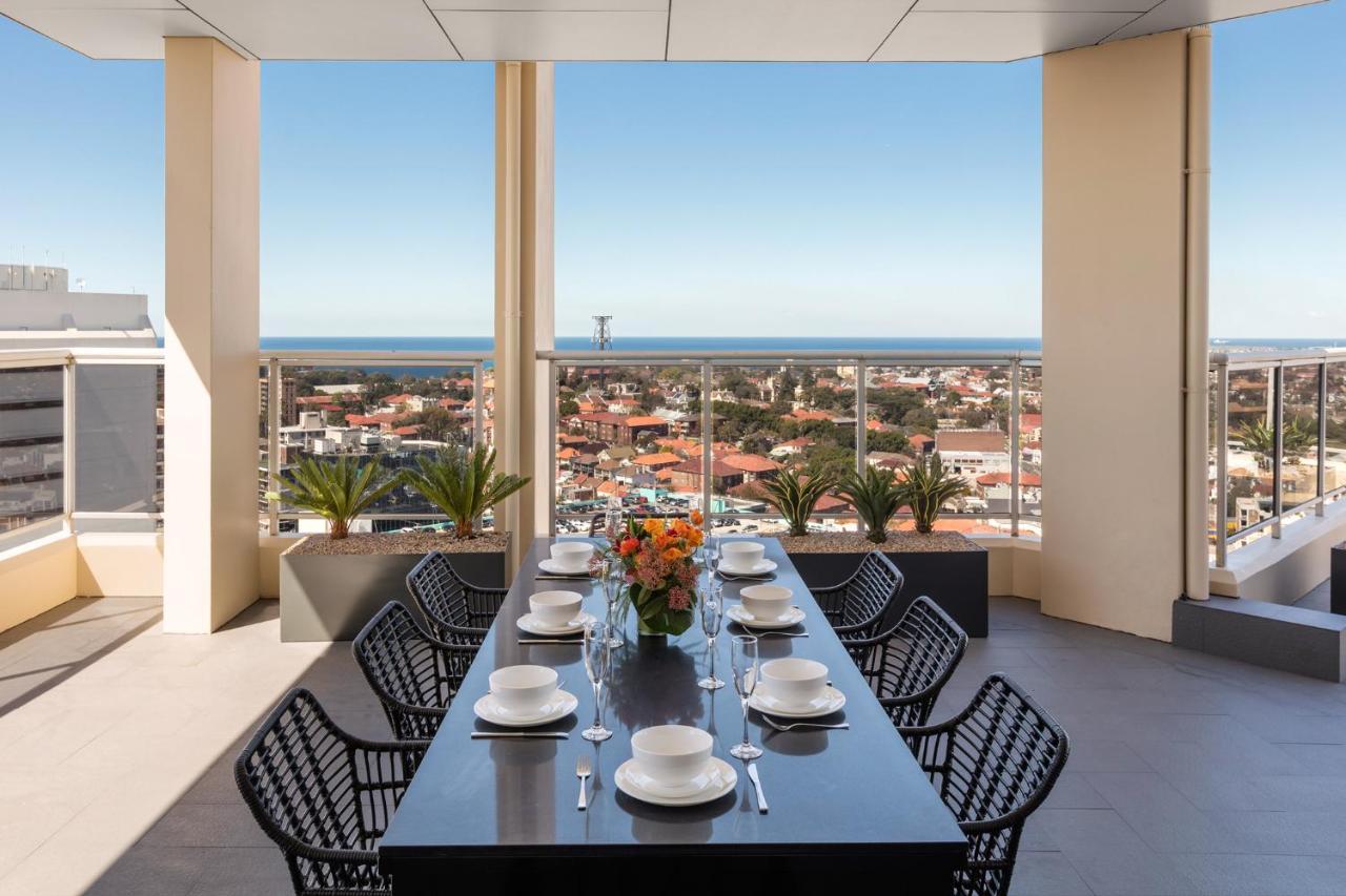 Meriton Suites Bondi Junction Sydney Bagian luar foto The photo shows a modern outdoor dining area situated on a balcony or terrace with a stunning view. There is a long rectangular table set with plates, glasses, and cutlery, ready for a meal. The table is surrounded by sleek, black wicker chairs. In t