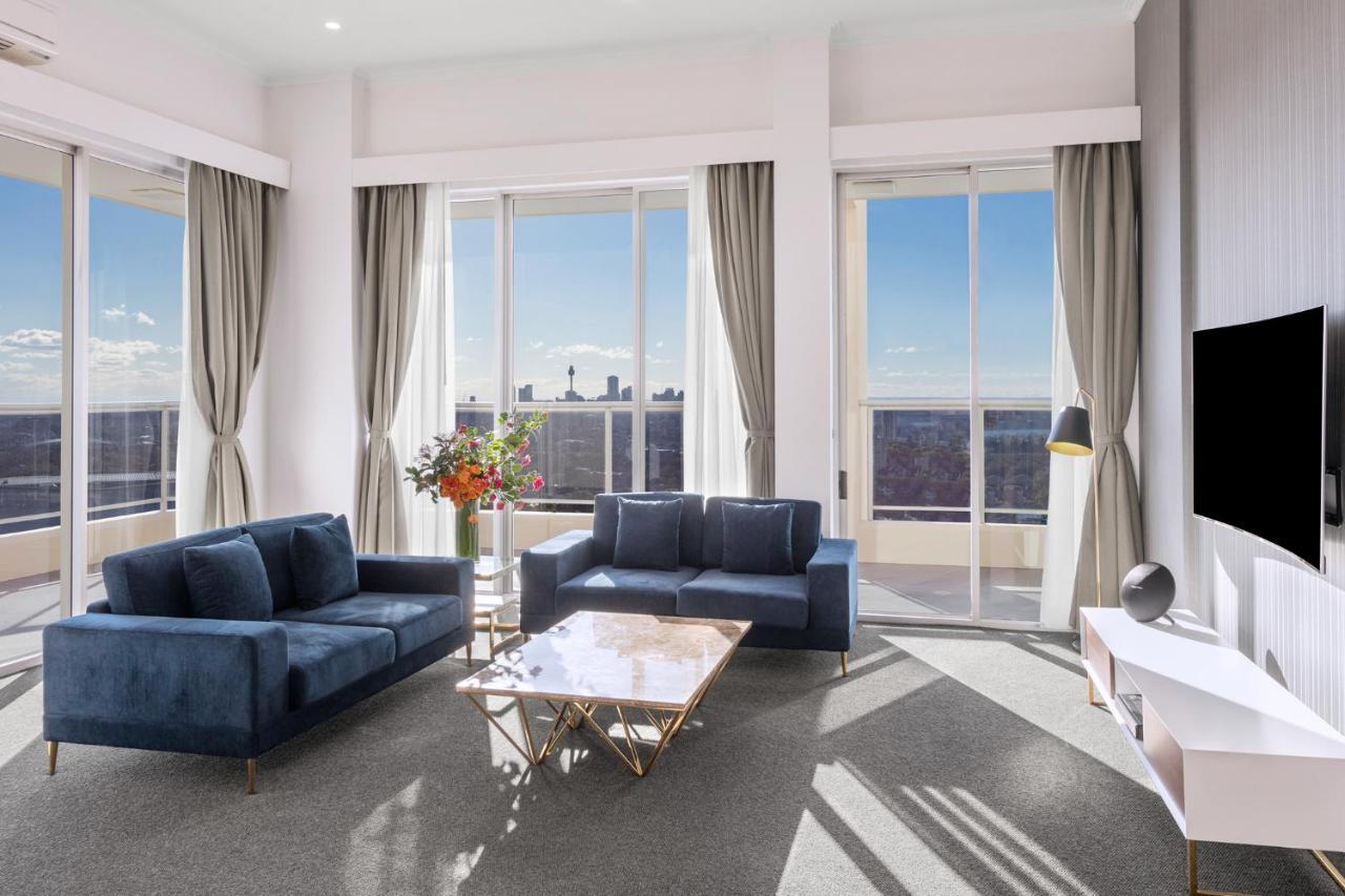 Meriton Suites Bondi Junction Sydney Bagian luar foto The photo shows a modern living room with large windows that allow natural light to fill the space. There are two blue sofas arranged around a coffee table with a glass top and gold legs. A floral arrangement adds a touch of color, placed on a nearby