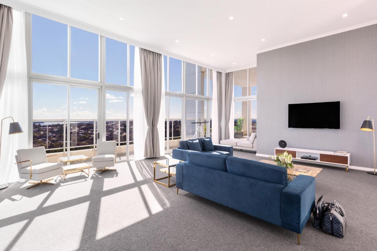 Meriton Suites Bondi Junction Sydney Bagian luar foto The photo shows a bright and modern living room with large floor-to-ceiling windows that provide a view of the outdoors. The space features a blue sofa in the center, complemented by light-colored armchairs nearby. There are light curtains hanging by