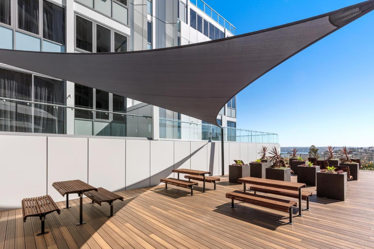 Meriton Suites Bondi Junction Sydney Bagian luar foto The photo shows a modern outdoor terrace area on a building. It features several wooden benches arranged on a wooden deck. There are large planters with greenery, adding some decorative elements. A large triangular gray shade sail is positioned above