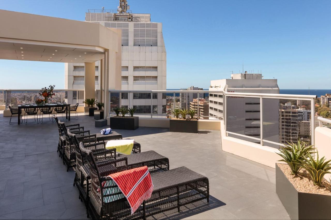 Meriton Suites Bondi Junction Sydney Bagian luar foto The photo shows a rooftop terrace with a modern design. There are several outdoor seating arrangements featuring black wicker lounge chairs. Some of the chairs have colorful blankets draped over them. The terrace is surrounded by a railing and has po