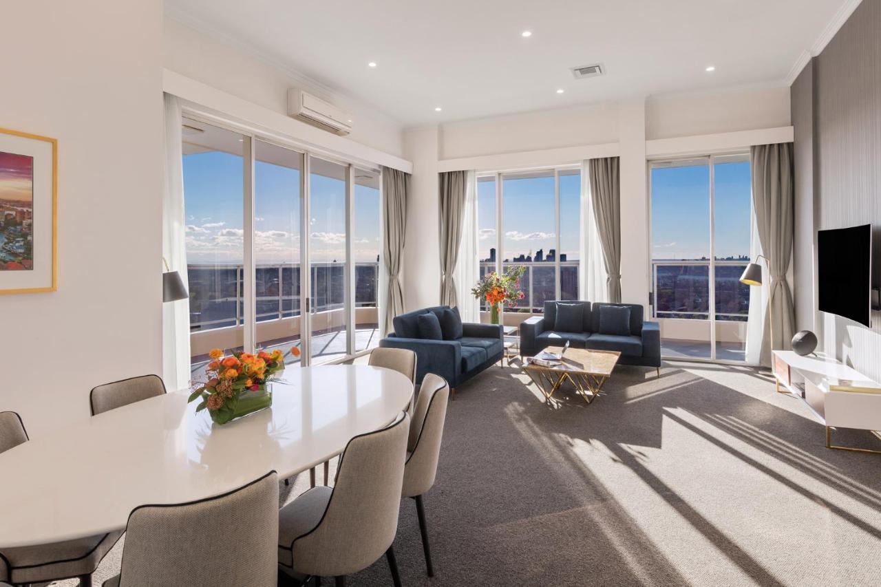 Meriton Suites Bondi Junction Sydney Bagian luar foto The photo shows a modern and stylish living space with large windows allowing natural light to fill the room. In the foreground, there is a round dining table with several chairs around it, adorned with a floral centerpiece. The seating area features