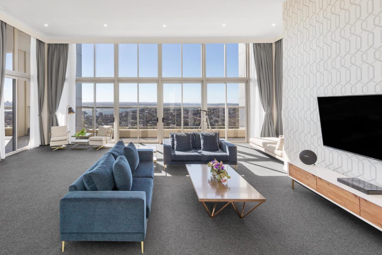 Meriton Suites Bondi Junction Sydney Bagian luar foto The photo shows a modern and spacious living area. It features large windows that provide a panoramic view of the outside landscape and allow natural light to fill the room. The interior design includes a blue sectional sofa placed in the center, com