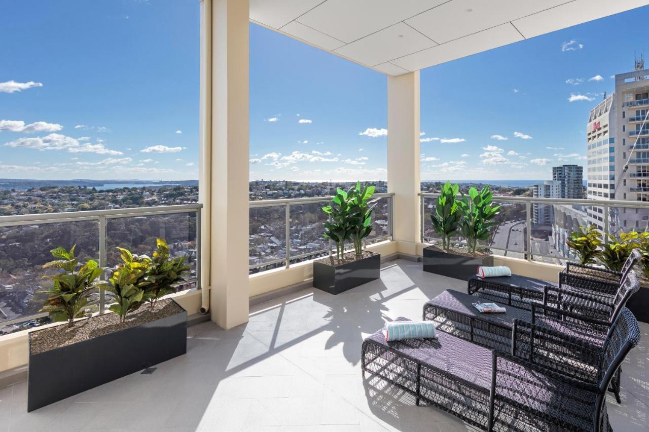 Meriton Suites Bondi Junction Sydney Bagian luar foto The photo shows a modern balcony or terrace area with a stunning view. There are several planters with green plants placed along the edge. The seating area features lounge chairs in a contemporary design, likely made of woven materials. The space app