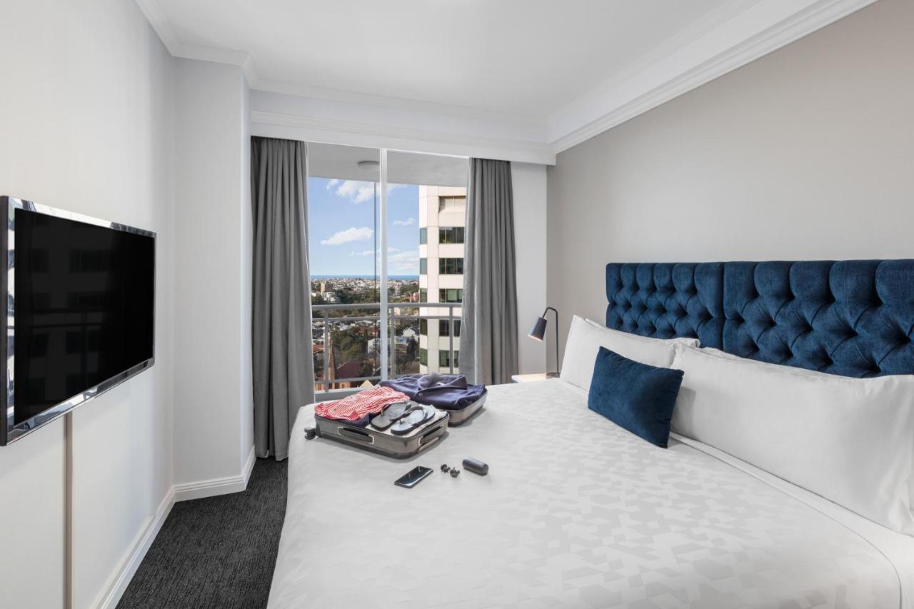 Meriton Suites Bondi Junction Sydney Bagian luar foto The photo shows a modern hotel room featuring a neatly made bed with a blue tufted headboard and a white duvet. There's a small decorative pillow on the bed, and a piece of luggage is placed at the foot with clothes partially unpacked. A television i