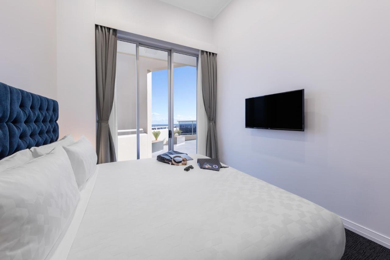 Meriton Suites Bondi Junction Sydney Bagian luar foto The photo shows a modern hotel room with a large, comfortable bed featuring a blue tufted headboard. There are light-colored walls, and the room is decorated simply. A flat-screen TV is mounted on the wall opposite the bed. The room has large glass d