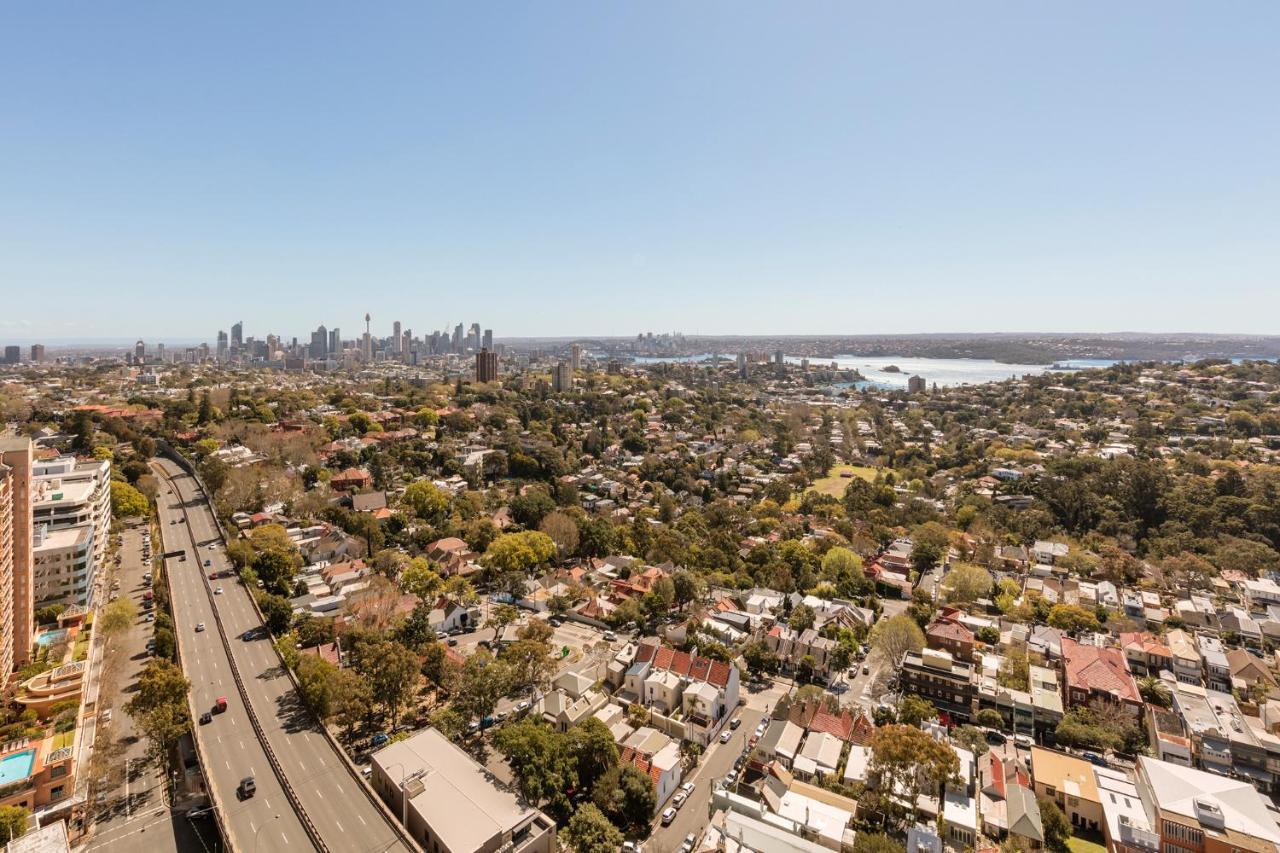Meriton Suites Bondi Junction Sydney Bagian luar foto The photo captures a panoramic view of a vibrant urban landscape. In the foreground, there are residential areas with various houses and greenery, showcasing a suburban environment. In the background, the skyline of a city can be seen, featuring tall