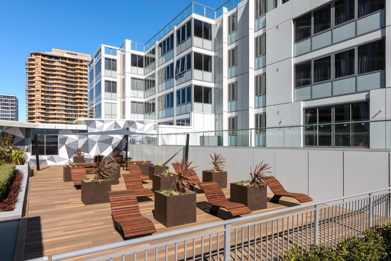 Meriton Suites Bondi Junction Sydney Bagian luar foto The photo shows a modern outdoor space featuring a wooden deck. There are several lounge chairs arranged around large planter boxes, which contain plants. In the background, there are contemporary residential buildings with large windows, and a geome