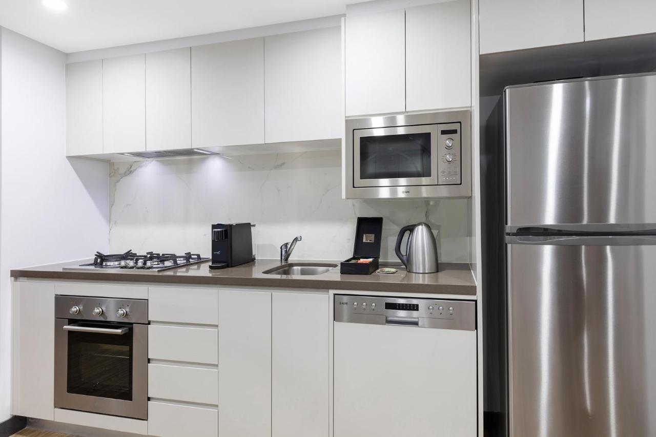 Meriton Suites Bondi Junction Sydney Bagian luar foto The photo shows a modern kitchen with a sleek, minimalist design. It features white cabinetry and a countertop that blends with the overall aesthetic. 

Key elements include:

- A stainless steel refrigerator on the right.
- An oven with a gas cookto