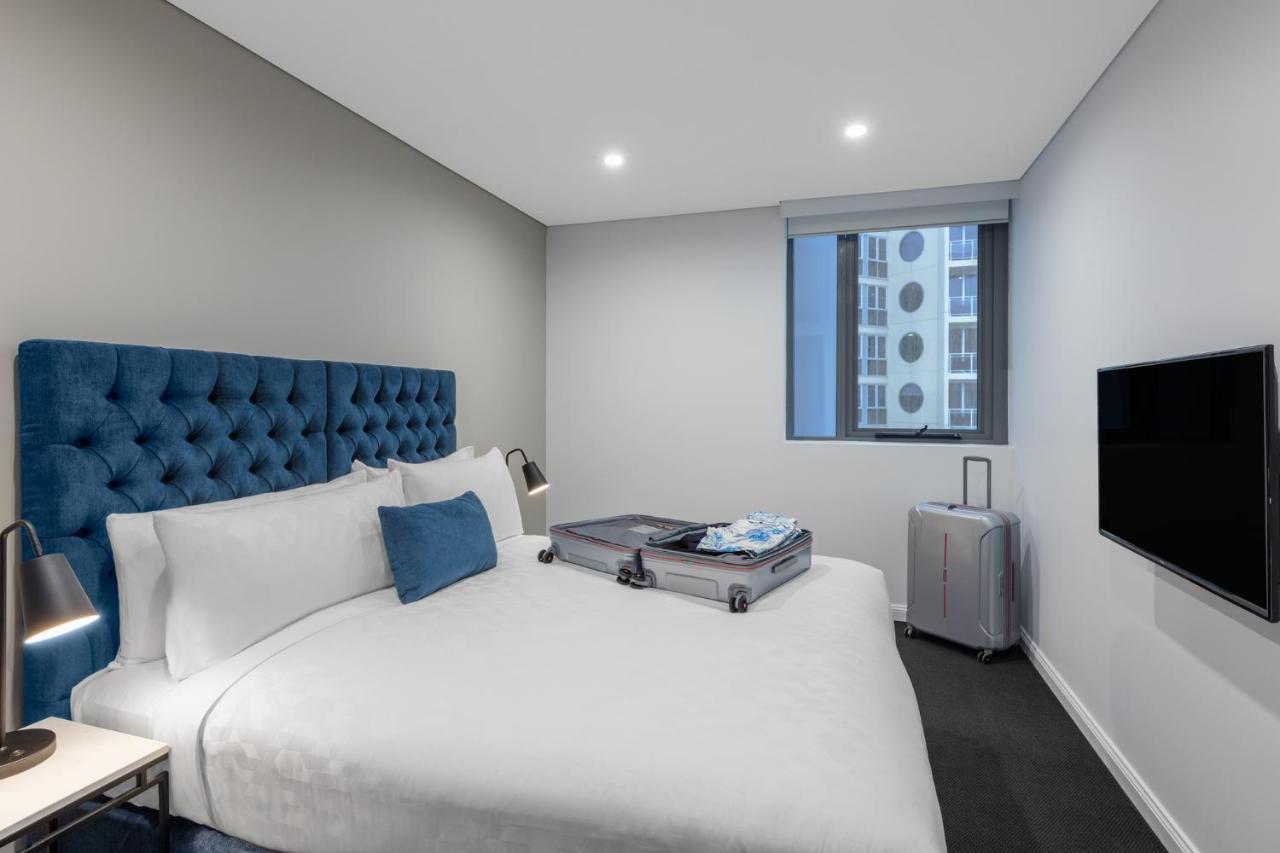 Meriton Suites Bondi Junction Sydney Bagian luar foto The photo shows a modern hotel room featuring a neatly made bed with a blue tufted headboard. There is a small blue decorative pillow on the bed, and the bedding is white. Next to the bed, there is a nightstand with a lamp. A suitcase is partially op