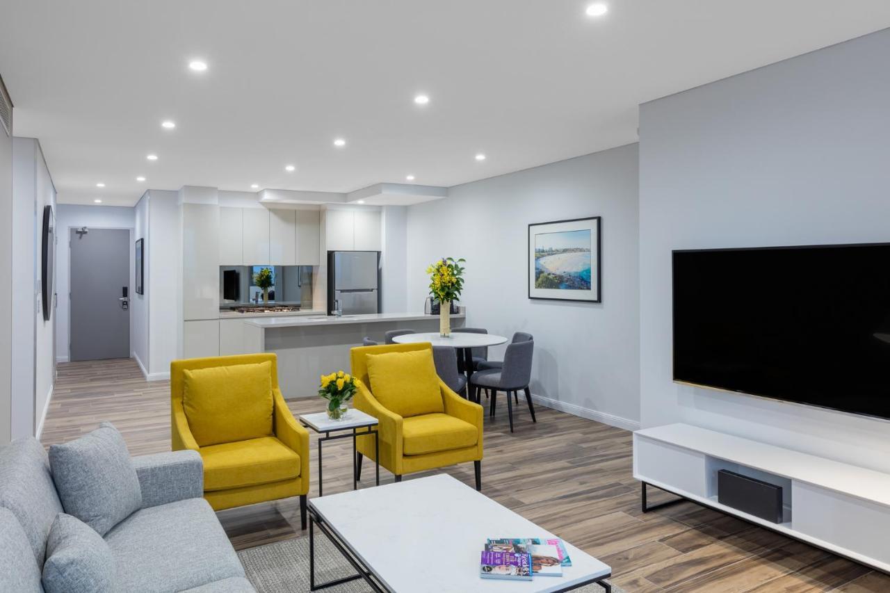 Meriton Suites Bondi Junction Sydney Bagian luar foto The photo shows a modern living space that includes a cozy sitting area and a dining area. The sitting area features two bright yellow chairs and a light gray sofa, arranged around a coffee table. There are decorative items on the table, including fl