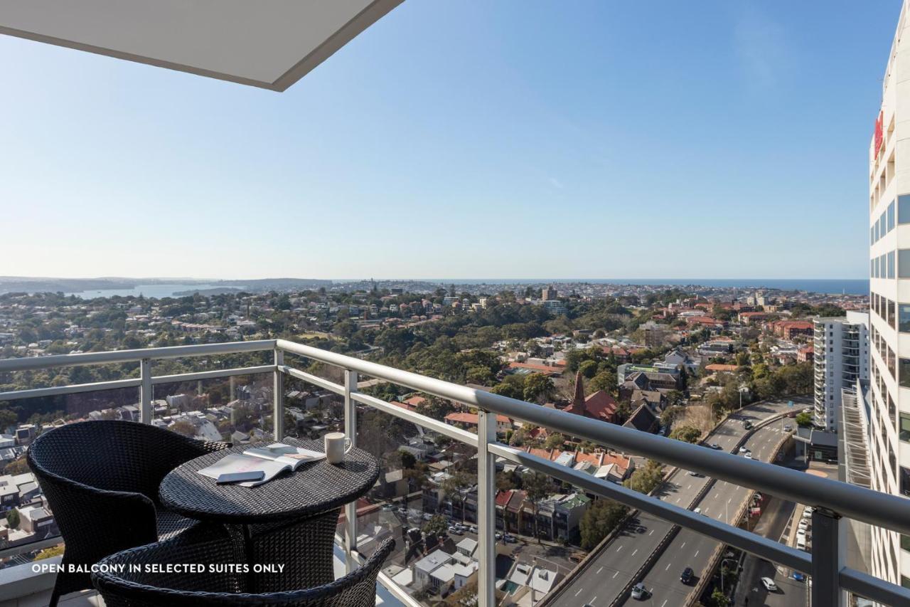 Meriton Suites Bondi Junction Sydney Bagian luar foto The photo shows a balcony with a view over a cityscape that includes buildings and greenery, extending towards the horizon where the sea is visible. The balcony features a small round table and two black chairs, suggesting a space for relaxation or d
