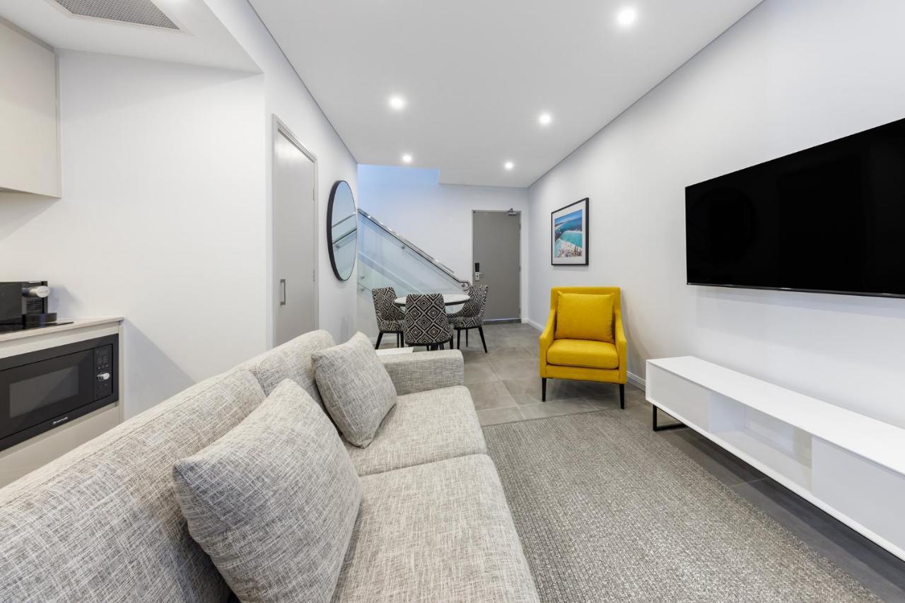 Meriton Suites Bondi Junction Sydney Bagian luar foto The photo shows a modern and stylish living space. It features a light-colored sofa with cushions, positioned to face a wall-mounted flat-screen TV. There's a yellow accent chair nearby that adds a pop of color to the room. The décor is minimalistic,