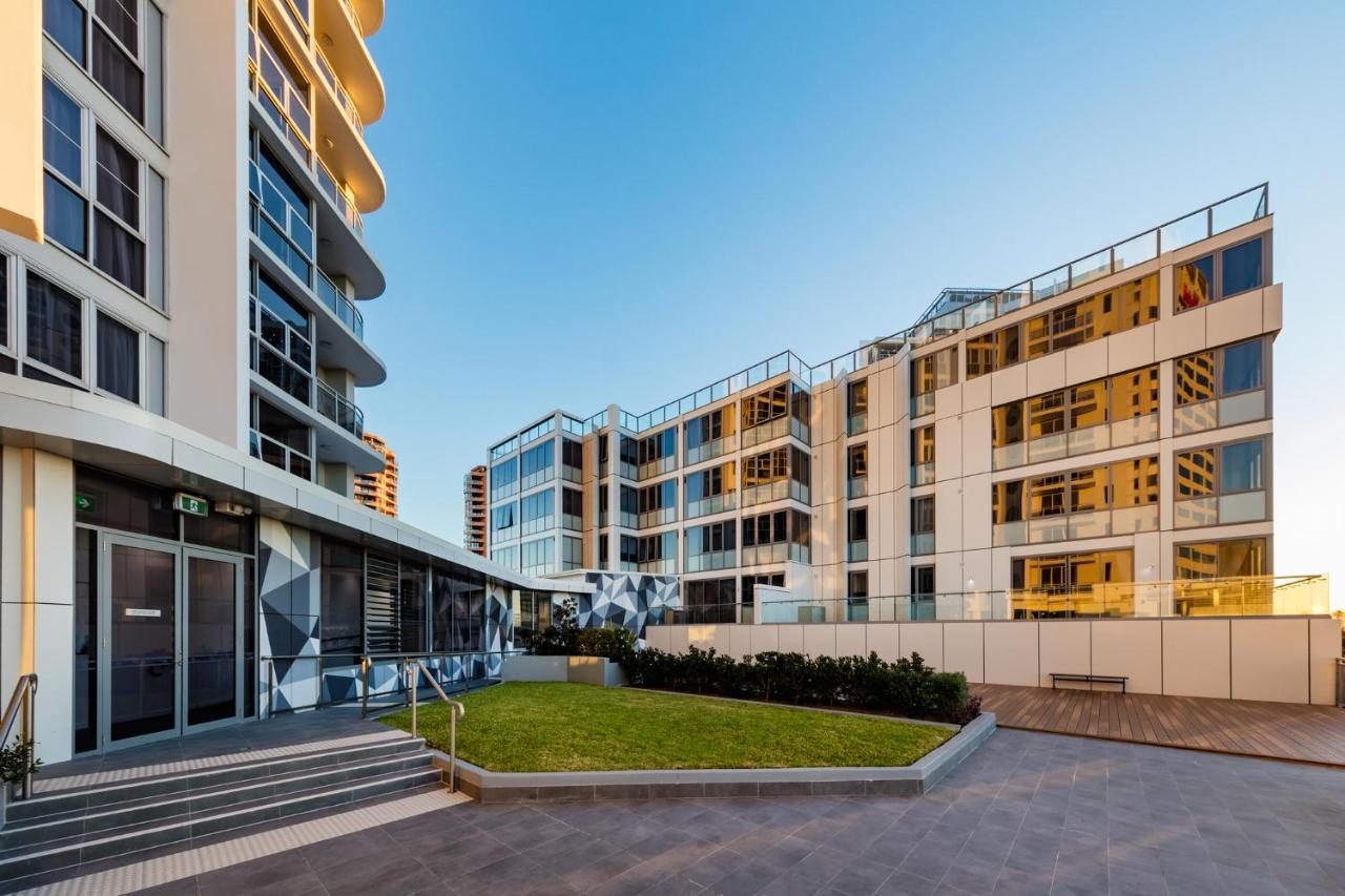 Meriton Suites Bondi Junction Sydney Bagian luar foto The photo showcases a modern residential area featuring contemporary buildings. In the foreground, there is a landscaped green space surrounded by well-designed architecture. The buildings have large glass windows, reflecting the light, and feature a