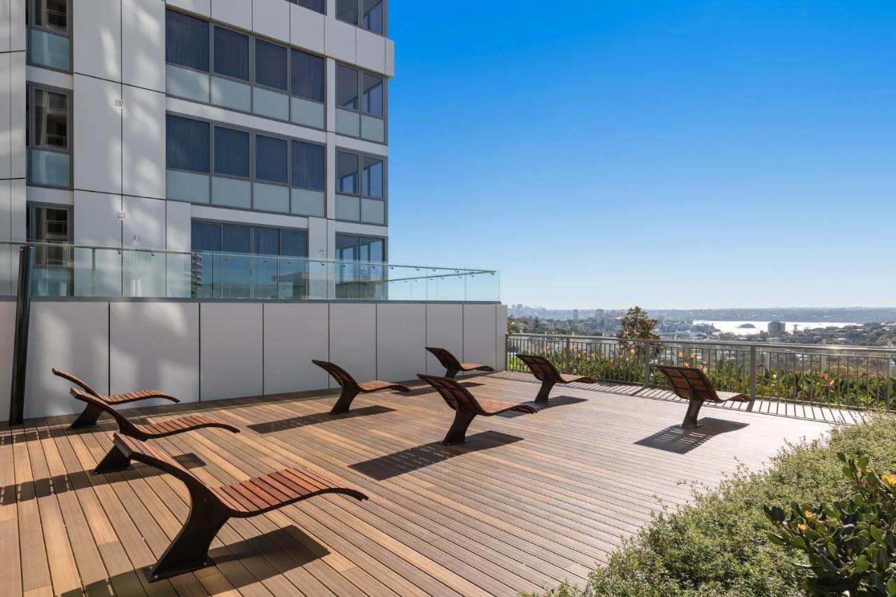Meriton Suites Bondi Junction Sydney Bagian luar foto The photo shows a rooftop or outdoor area with a wooden deck. There are several modern lounge chairs arranged neatly, facing a clear sky and a panoramic view of the surrounding landscape. In the background, there is a tall building with large windows