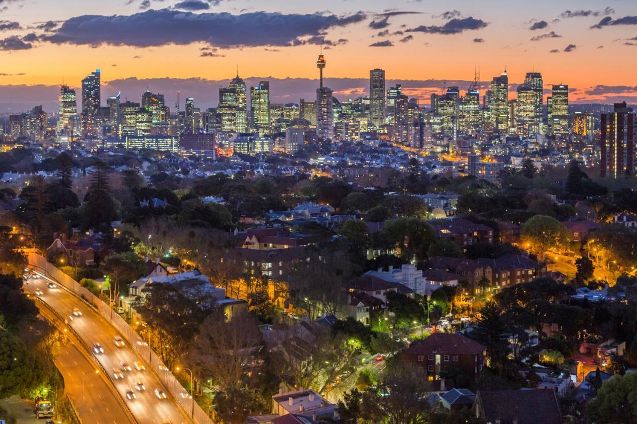 Meriton Suites Bondi Junction Sydney Bagian luar foto The photo captures a stunning city skyline at dusk, featuring towering skyscrapers adorned with lights against a colorful sunset backdrop. In the foreground, a green area with trees and houses is visible, while a road with cars can be seen winding th