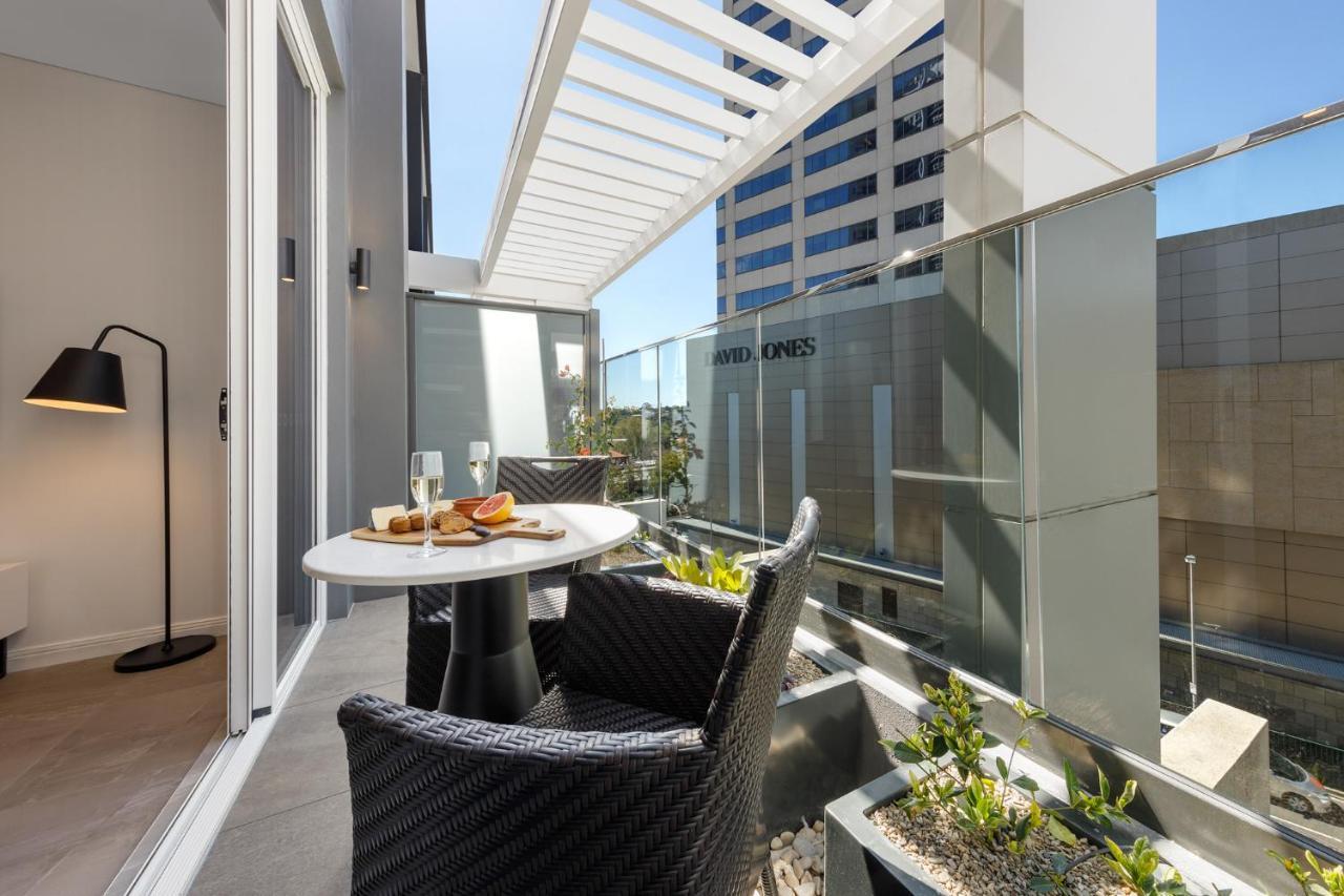 Meriton Suites Bondi Junction Sydney Bagian luar foto The photo shows a modern balcony area with glass railings. It features a small round table set for two, with a plate of food and drinks. There are two stylish wicker chairs positioned around the table. The space is bright and airy, with sunlight stre