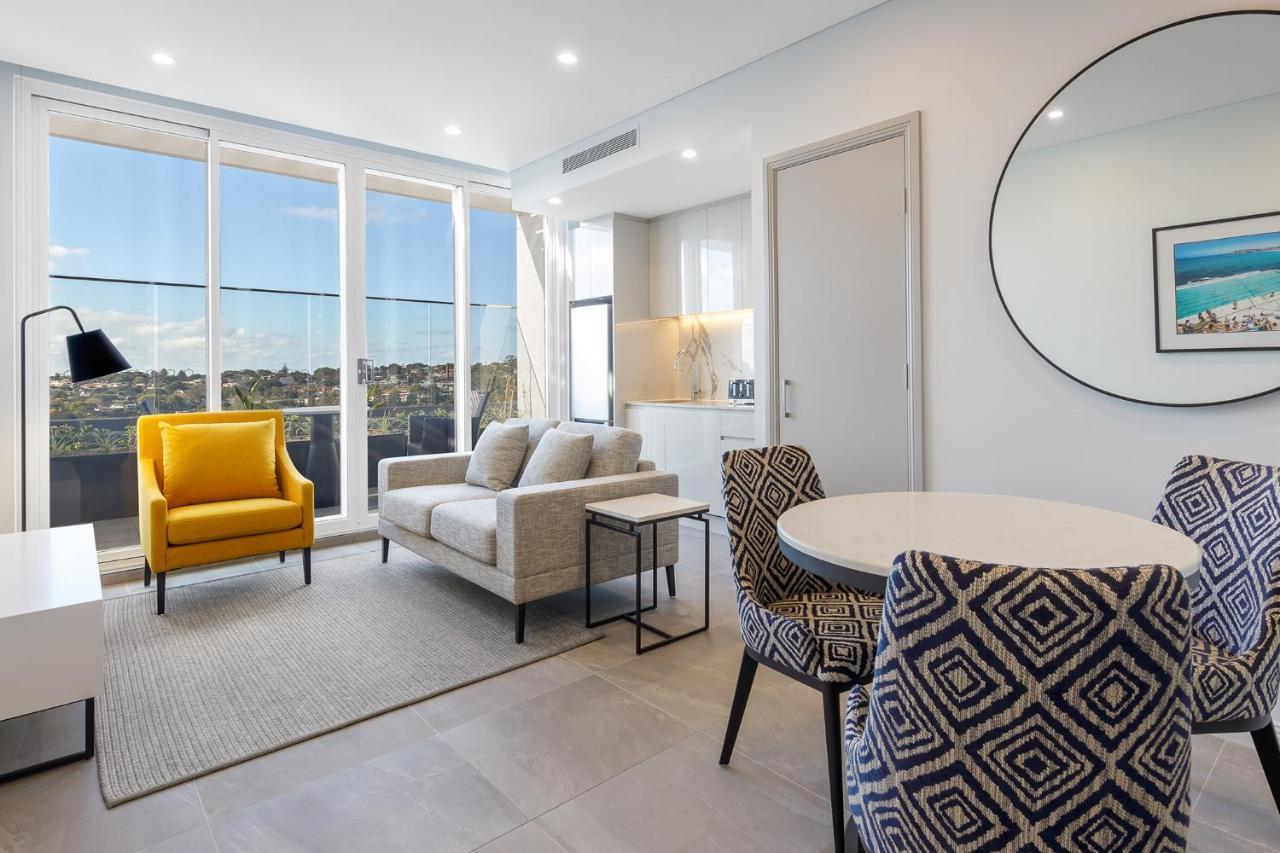 Meriton Suites Bondi Junction Sydney Bagian luar foto The photo shows a modern and stylish living area. It features a comfortable sofa in a neutral color, accompanied by a bright yellow armchair that adds a pop of color. There's a round dining table with patterned chairs nearby. Large windows allow plen