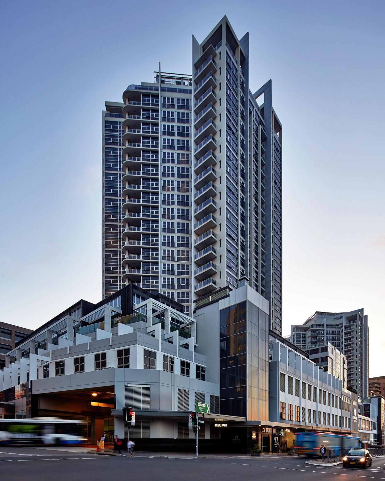 Meriton Suites Bondi Junction Sydney Bagian luar foto The image shows a modern high-rise building with a sleek design. The structure features a mix of glass and concrete elements, with multiple levels showcasing balconies. The building appears to be part of an urban environment, likely containing reside