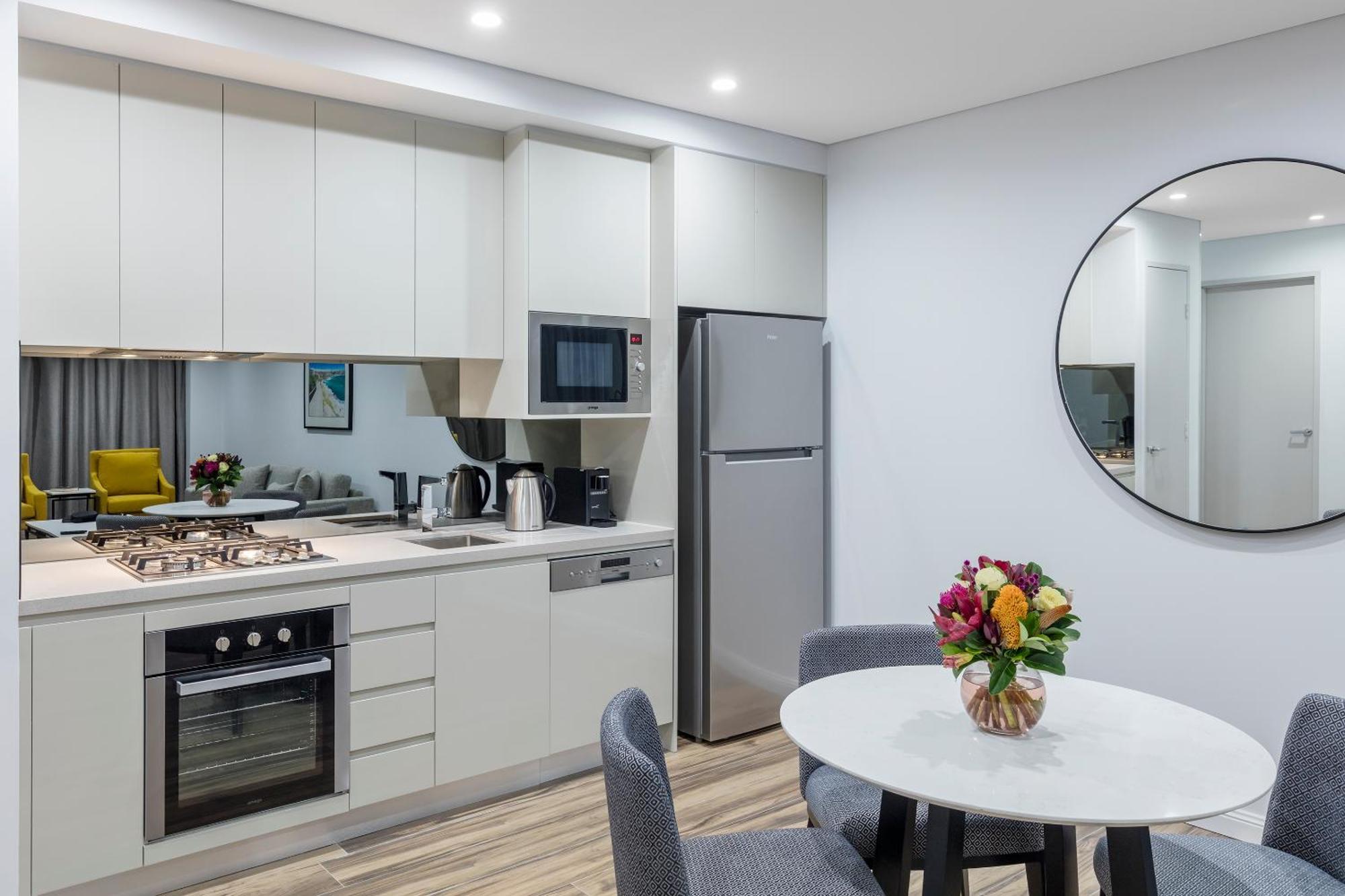 Meriton Suites Bondi Junction Sydney Bagian luar foto The photo shows a modern kitchen and dining area. The kitchen features light-colored cabinetry with a built-in oven and stovetop, a microwave, and a silver refrigerator. There is a kettle and a coffee maker on the countertop. In the dining area, ther